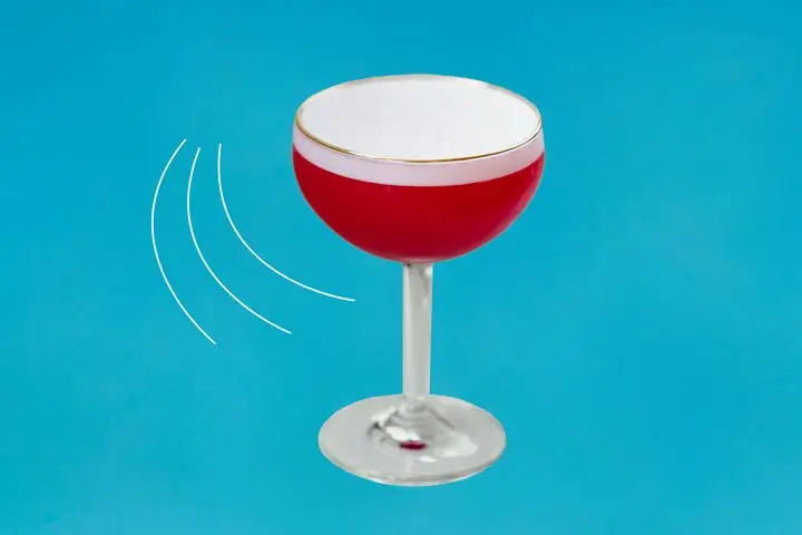 On a blue background, an elegant coupe holds a vivid and opaque red drink. A thick layer of white foam rests atop the drink