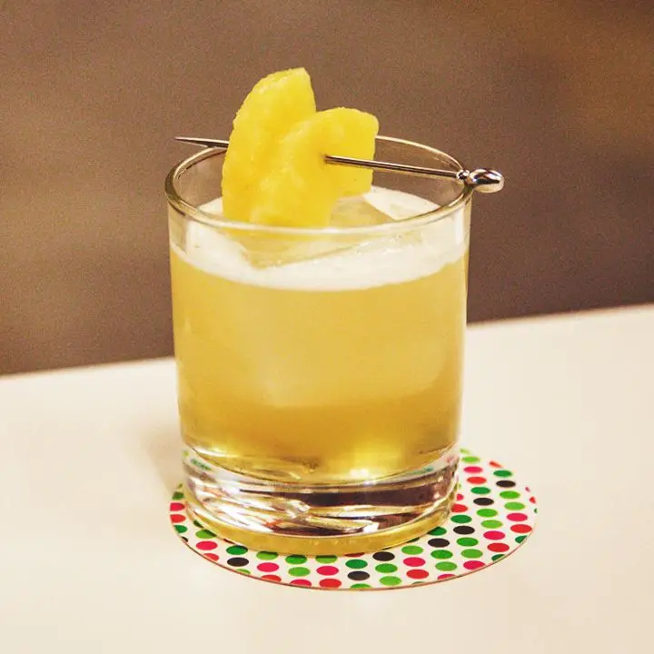 A shaken yellow-hued cocktail in a rocks glass garnished with two manicured slices of pineapple
