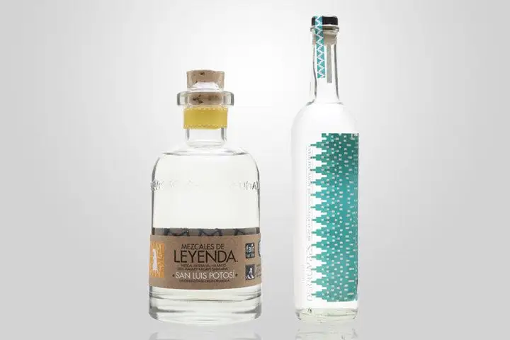 Two different mezcal bottles, Mezcales de Leyenda and Derrumbes, pictured next to one another against a gray gradient background