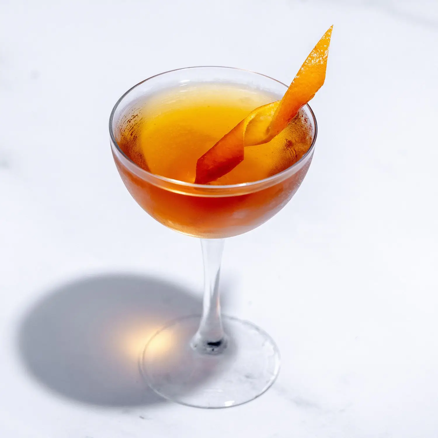 Overhead-angled view of a Martinez cocktail in a coupe class on white marbled background, with orange twist along the rim