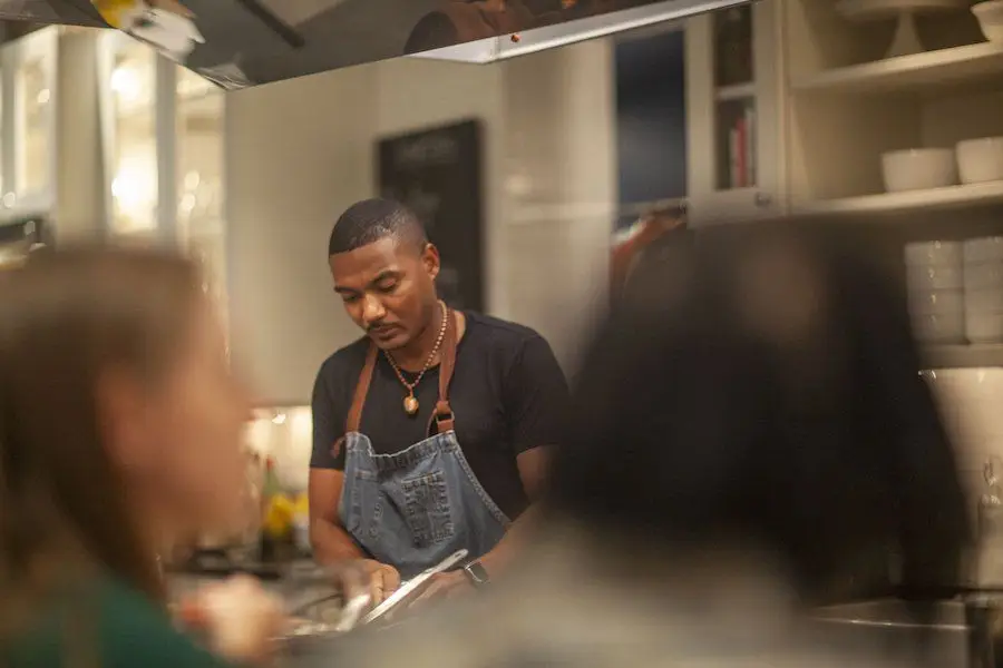 A chef in a black T-shirt and a denim apron preparing dinner for a small group of guests