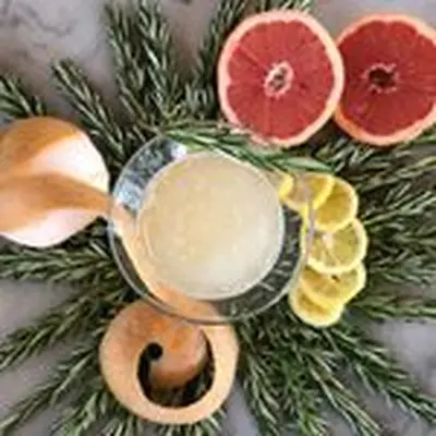 A pale cocktail in a Martini glass is centered in a top-down photo. Surrounding it is a wreath of rosemary, as well as a partially peeled grapefruit, a lemon sliced into rings, and a halved grapefruit. The background is marble.