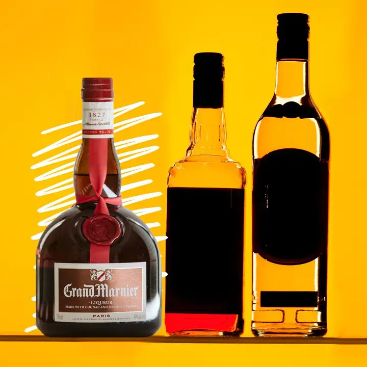A stout Grand Marnier bottle sits on a yellow background. Next to it are other liquor bottles, their labels in shadow