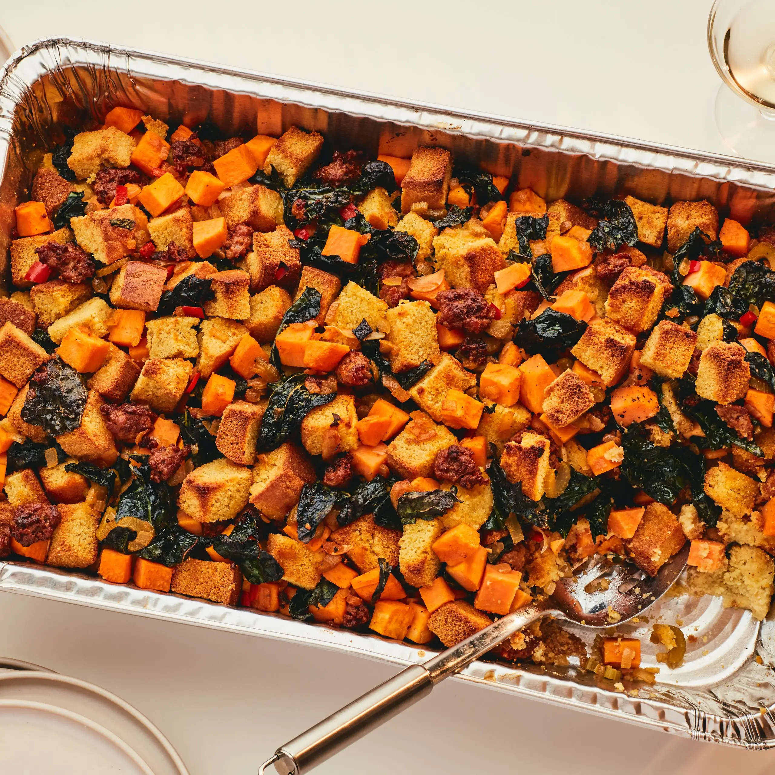 Large disposable aluminum platter of sweet potato and kale stuffing with serving spoon and wine on the side.